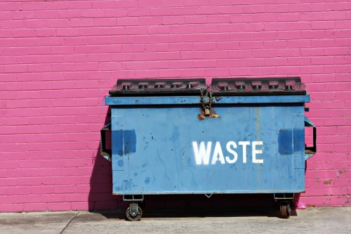 A large blue industrial waste skip with the word 'WASTE' painted in white on the front, positioned on a concrete pavement against a vibrant pink brick wall. The skip has a hinged lid with multiple ventilation holes, and appears weathered with visible dirt and rust marks around the latch area. It is mounted on black wheels, allowing mobility, and is situated in an outdoor urban environment, possibly near a commercial property in Neasden. The surrounding area is clean, with no other objects or debris visible nearby, highlighting its role in waste collection services provided by companies like Commercial Waste Neasden. The lighting suggests daylight, casting shadows on the ground and emphasizing the textures of the bricks and the metal surface of the skip, conveying a practical and straightforward waste management scene suitable for a professional rubbish removal context in the local area.