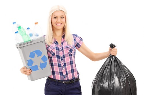 A young woman with long blonde hair and a cheerful expression is standing against a plain white background, holding a grey recycling bin filled with various plastic bottles and containers on her left side, and a tightly tied black plastic rubbish bag in her right hand. She is dressed in a casual short-sleeved, pink and blue checked shirt and dark trousers. The recycling bin features a prominent blue recycling symbol on the front. The image appears to be a promotional photograph, with the woman presenting waste collection and recycling services, which could relate to rubbish removal companies such as Commercial Waste Neasden. The scene emphasizes organized waste disposal, with a focus on plastic recyclables and general rubbish collection, typical of a domestic or small commercial waste management context, set against a clean background to highlight environmental responsibility and waste handling.