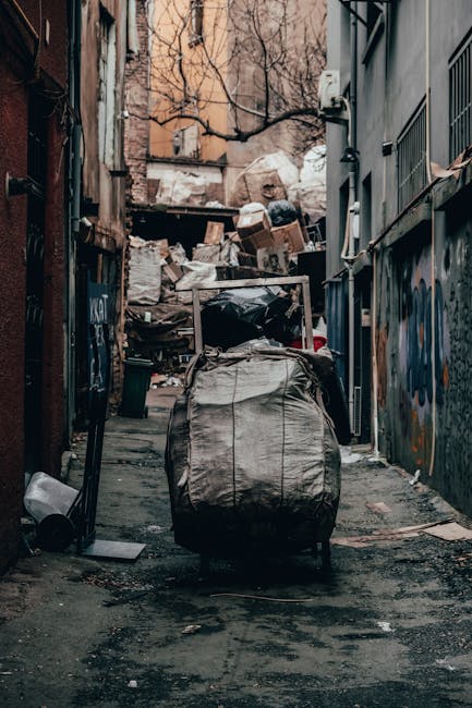 A close-up view of an overflowing pile of mixed waste materials situated on a gravel surface outdoors. The refuse includes black plastic garbage bags, some torn open revealing crumpled paper, plastic 