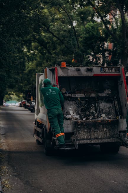 A worker dressed in green high-visibility work overalls and a matching helmet is standing next to the open rear loading bay of a commercial rubbish collection vehicle on a residential street. The vehi