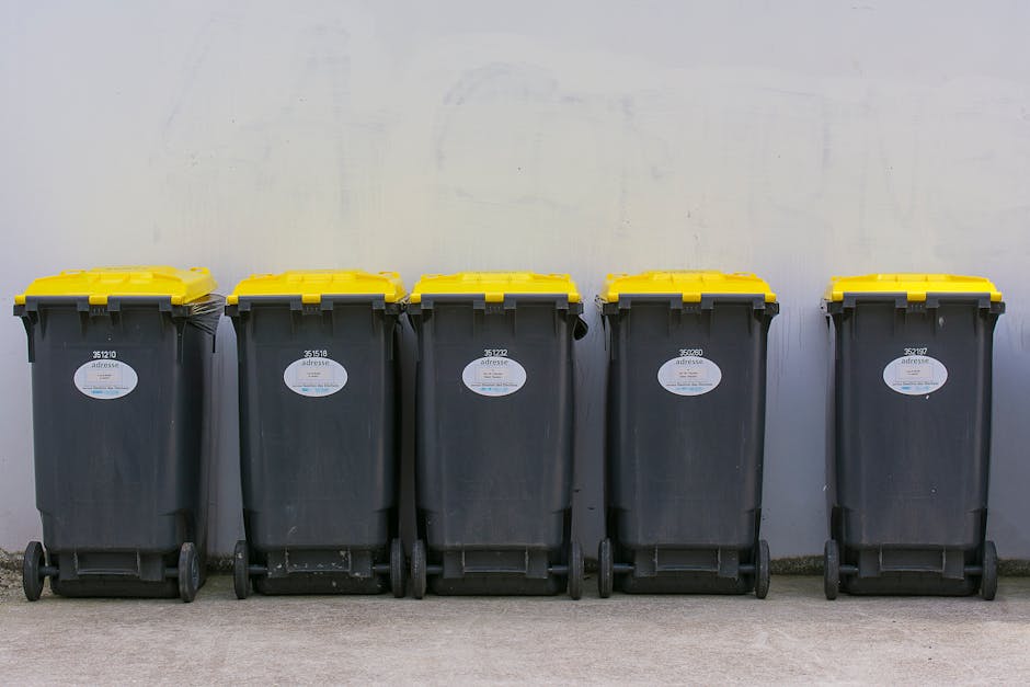 A worker dressed in green high-visibility work overalls and a matching helmet is standing next to the open rear loading bay of a commercial rubbish collection vehicle on a residential street. The vehi
